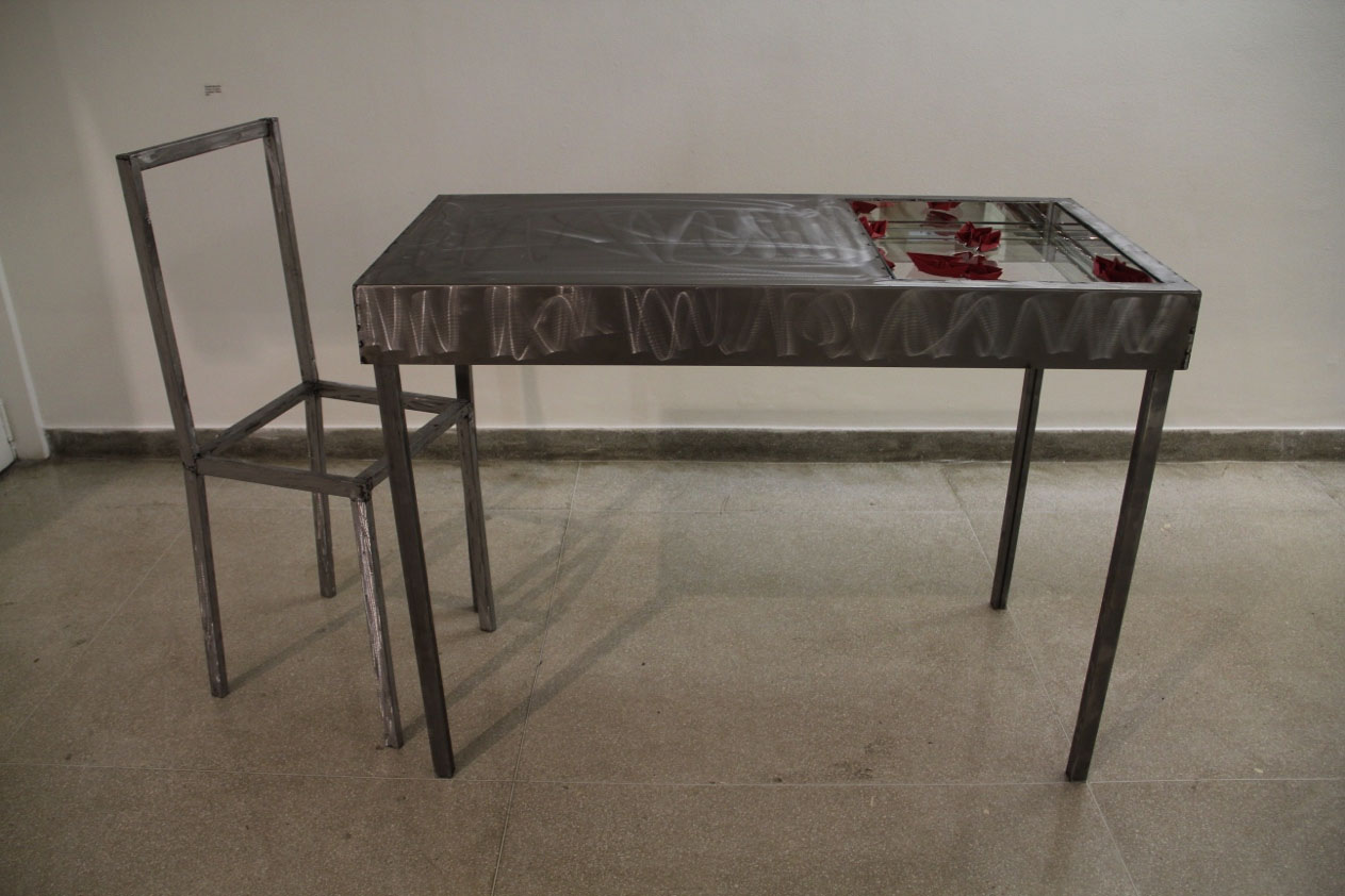Un chaise sans assise fait face à une table en métal dont la moitié est creusée et recouverte d'un miroir. A la surface du miroir des bateaux en origami rouge sang.